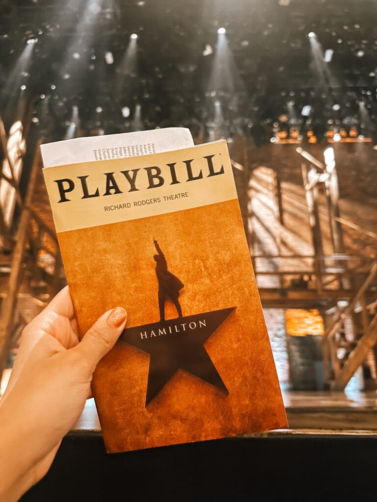 A hand holding a “Hamilton” Playbill in front of the stage at the Richard Rodgers Theatre. Warm lighting shines down on the wooden set pieces in the background.