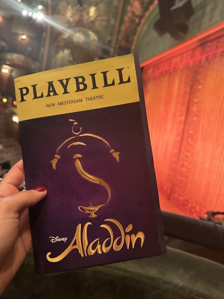A hand with red nail polish holds up a Playbill for Disney’s “Aladdin” at the New Amsterdam Theatre. The Playbill has a purple cover with a golden magic lamp and swirling genie silhouette. In the background, the theater’s ornate interior and glowing red curtain are softly blurred.