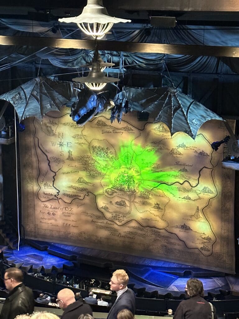 A wide view of the “Wicked” theater stage before the show, featuring a large map of Oz illuminated with green light and a mechanical dragon hanging overhead. Audience members are taking their seats below.